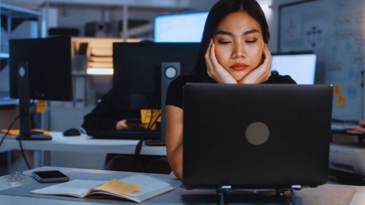 Signs You’re Ready for a Career Change and How Career Coaching in Temecula, CA Can Help You Find Fulfillment 2 A tired office employee sits at a desk resting their head in their hands behind a laptop, with an open notebook, smartphone, and other computer workstations visible in a dimly lit workspace, suggesting boredom or burnout at work.