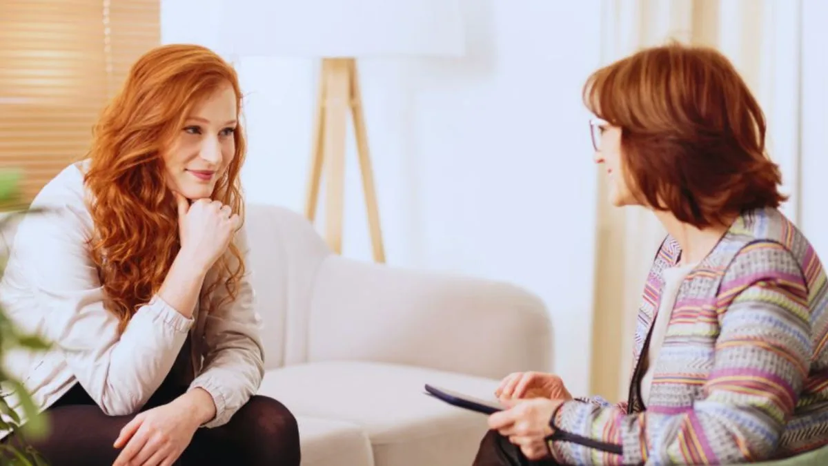 Two women sit facing each other in a light, modern office, with one appearing distressed and the other holding a tablet and listening attentively, depicting a supportive career coaching session.