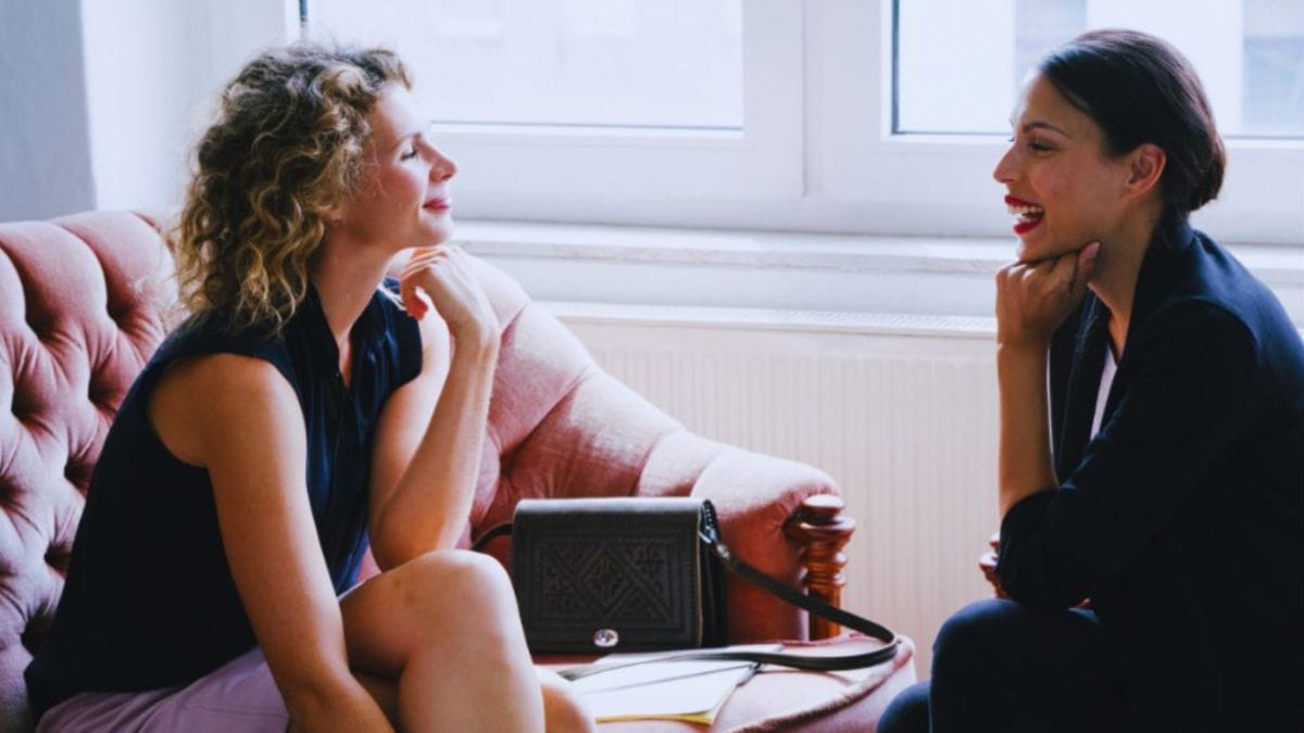 Two women sit facing each other on a pink sofa in a bright office, engaged in a thoughtful one-on-one conversation, illustrating a supportive career coaching session in Menifee, CA.