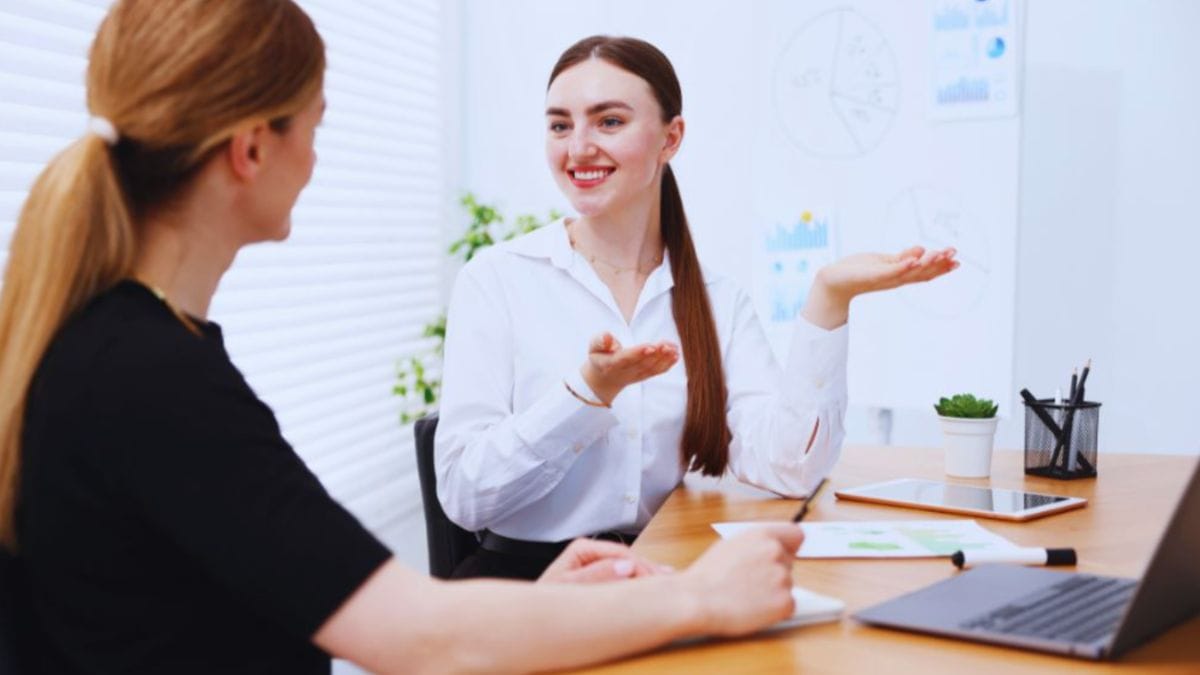 Two professional women sit across a desk in a bright office, with one coach gesturing as she explains career strategies using charts and a laptop, representing career coaching services.